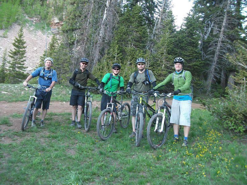 A group of five individuals stands on a grassy trail surrounded by tall trees, each holding a mountain bike. They are dressed in outdoor sports attire, including helmets and gloves, ready for biking. The setting suggests a recreational activity in a mountainous environment on a clear day. The Whole Enchilada mountain bike trail.