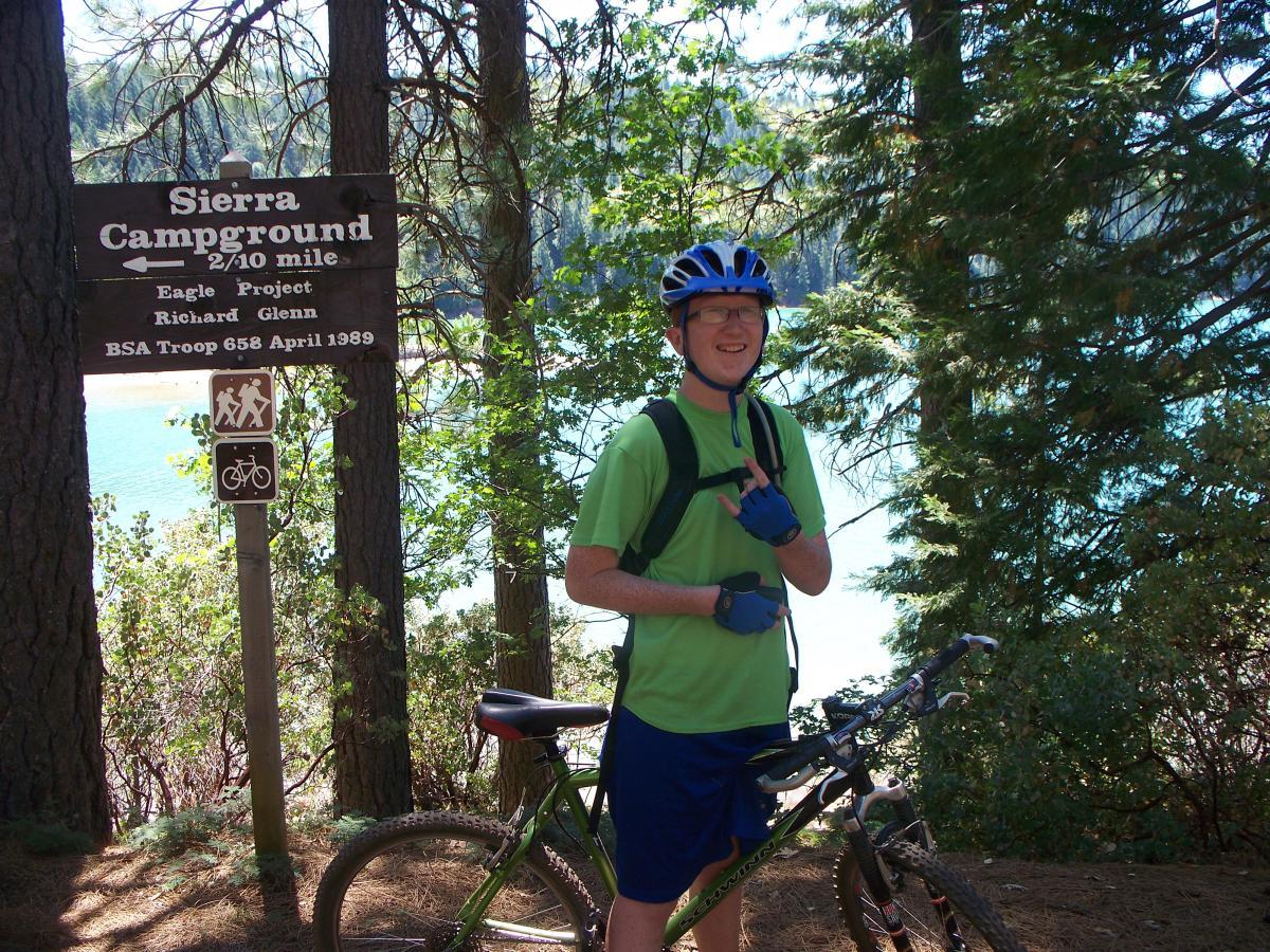 A young man wearing a green t-shirt, blue gloves, and a helmet stands next to a mountain bike in front of a sign for Sierra Campground. The sign indicates the campground is 2/10 mile away and displays symbols for hiking and biking. Tall trees and a lake can be seen in the background, suggesting a scenic outdoor setting. Sly Park mountain bike trail.