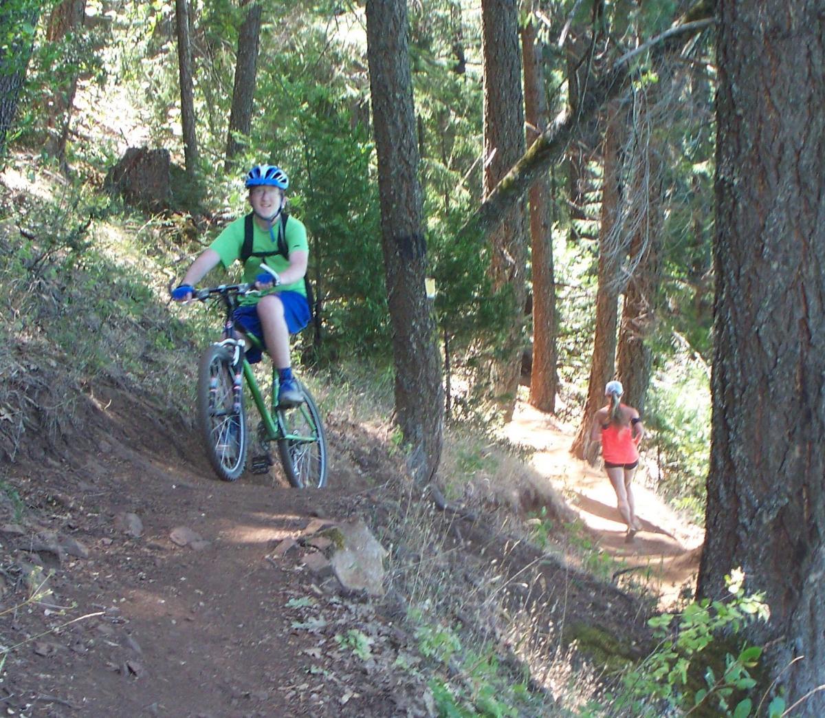 A young person riding a green mountain bike on a dirt trail surrounded by tall trees, while another person jogs on a nearby path. The scene captures an outdoor activity in a nature setting. Sly Park mountain bike trail.