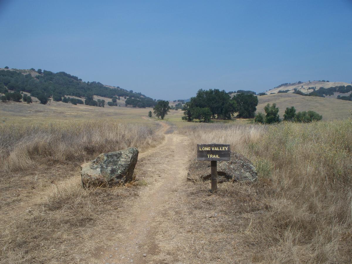 A dirt trail leading into a scenic valley, flanked by dry grass and large rocks. A wooden sign labeled "Long Valley Trail" is positioned at the trailhead, with rolling hills and trees visible in the background under a clear blue sky. Up Down Trail mountain bike trail.