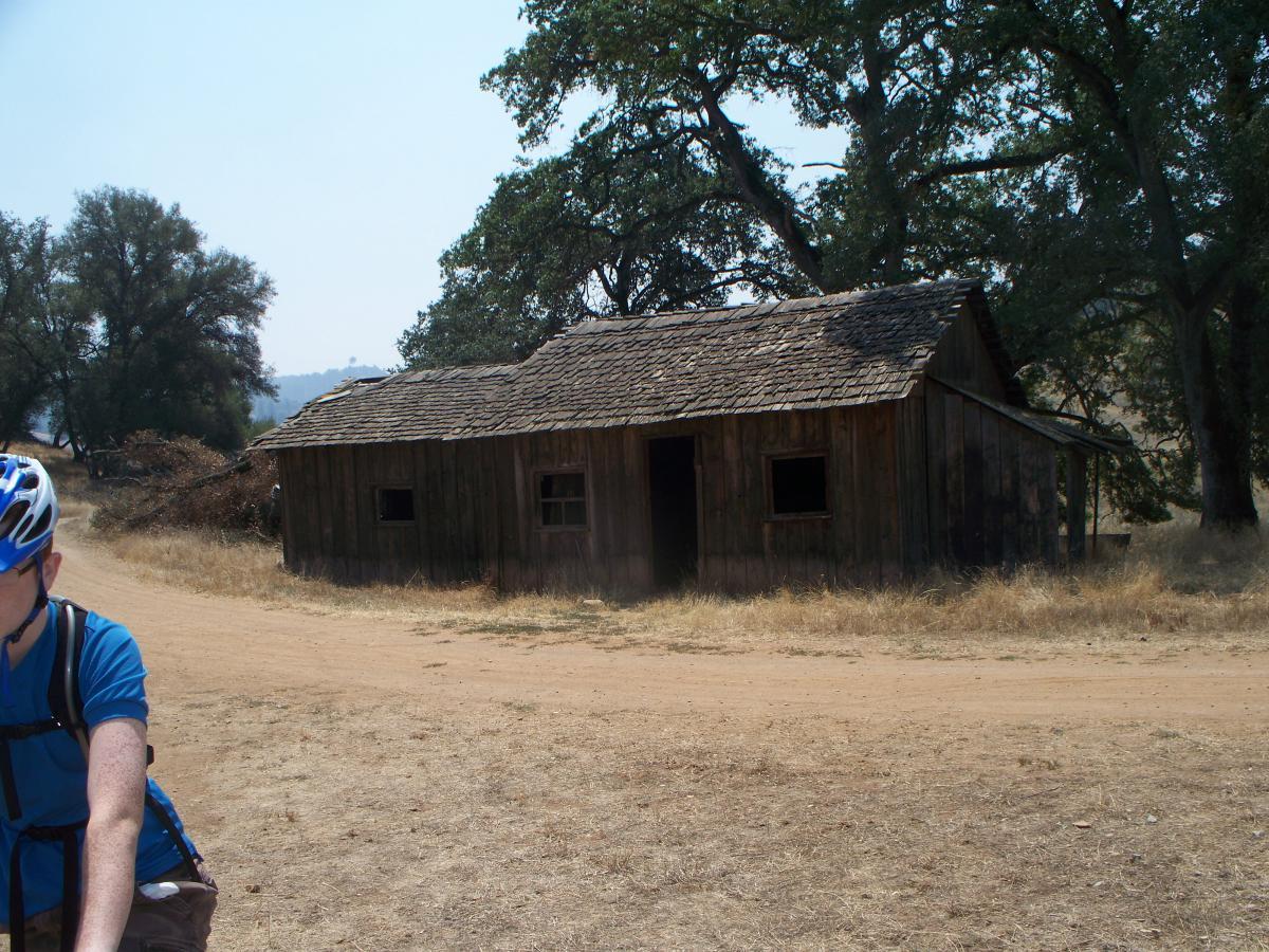 A rustic wooden cabin with a sloped roof, surrounded by dry grass and a dirt road. Sparse trees are visible in the background, contributing to a natural, rural landscape. A person in a blue shirt and helmet is partially visible in the foreground, engaging in an outdoor activity next to the cabin. Up Down Trail mountain bike trail.