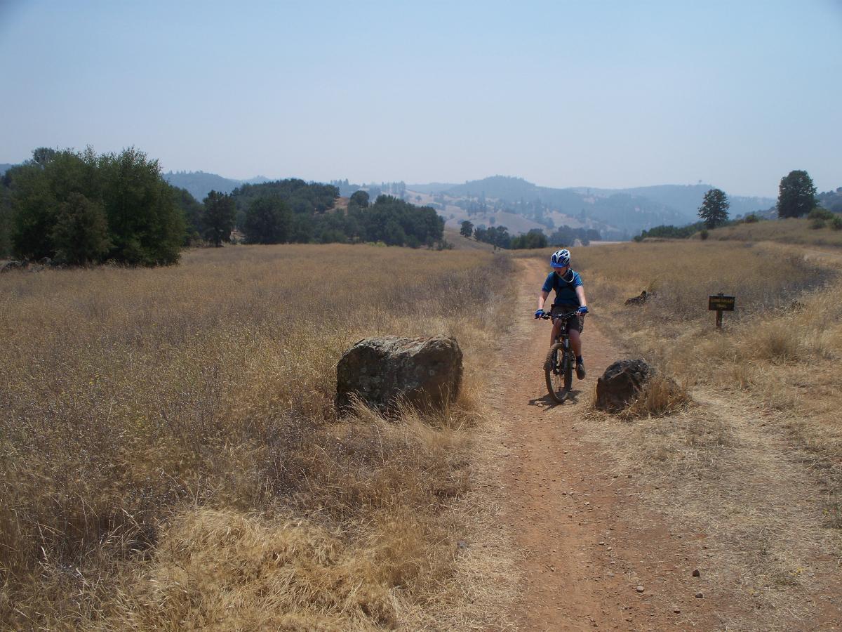 A person wearing a blue helmet and shirt rides a mountain bike along a dirt trail in a grassy, open area. There are large rocks on either side of the path, and the background features rolling hills and trees under a clear sky. A sign can be seen in the distance. Up Down Trail mountain bike trail.