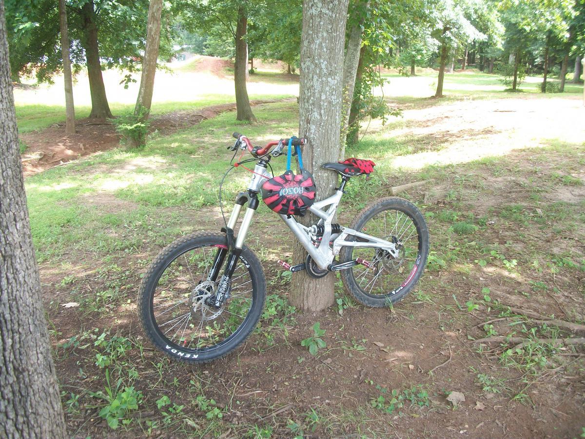 A mountain bike leaning against a tree in a wooded area, with a clear view of the bike's frame and tires. The ground is partially covered with grass and dirt, and there are several trees in the background, creating a natural, outdoor setting. Gateway Mountain Bike Park mountain bike trail.
