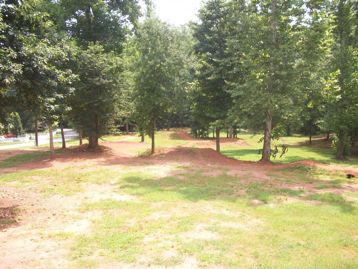 A sunny outdoor scene featuring a motocross track surrounded by trees. The ground is uneven with dirt mounds, indicating a course for biking or dirt biking, while green grass and patches of dirt are visible throughout the area. A road is visible in the background, suggesting a nearby pathway. Gateway Mountain Bike Park mountain bike trail.