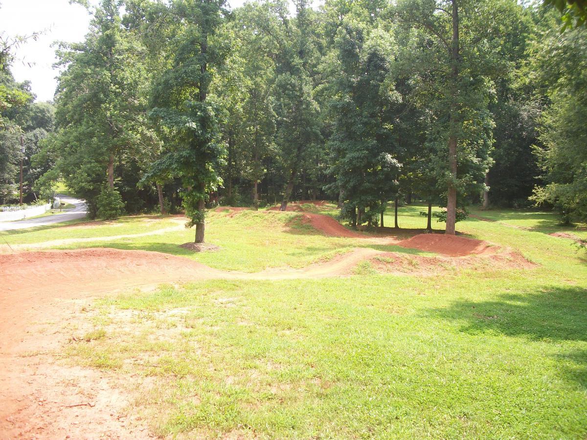 A view of a dirt track with gentle hills and turns, surrounded by lush greenery and trees. A road can be seen in the distance, with a clear blue sky above. The area appears to be recreational, suitable for outdoor activities such as biking or running. Gateway Mountain Bike Park mountain bike trail.