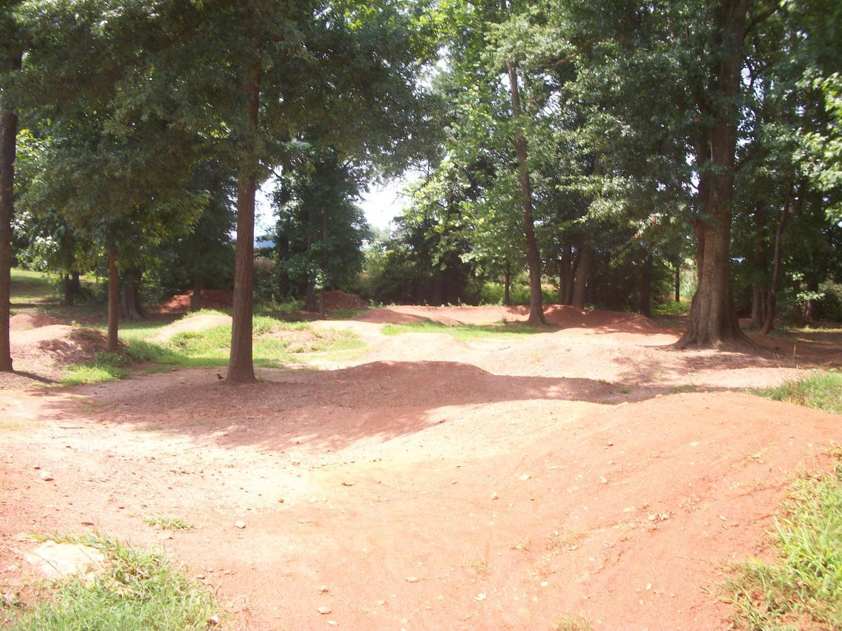 A dirt bike track surrounded by trees, featuring several dirt mounds and dips, set in a natural outdoor environment. The area is mostly covered in reddish soil with patches of grass visible. Gateway Mountain Bike Park mountain bike trail.