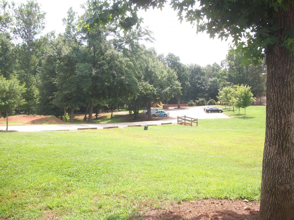 A lush, green park scene with a grassy foreground and a background of trees. A paved area is visible in the middle, featuring parked cars and a small playground area. The sunlight filters through the foliage, creating a serene outdoor environment. Gateway Mountain Bike Park mountain bike trail.