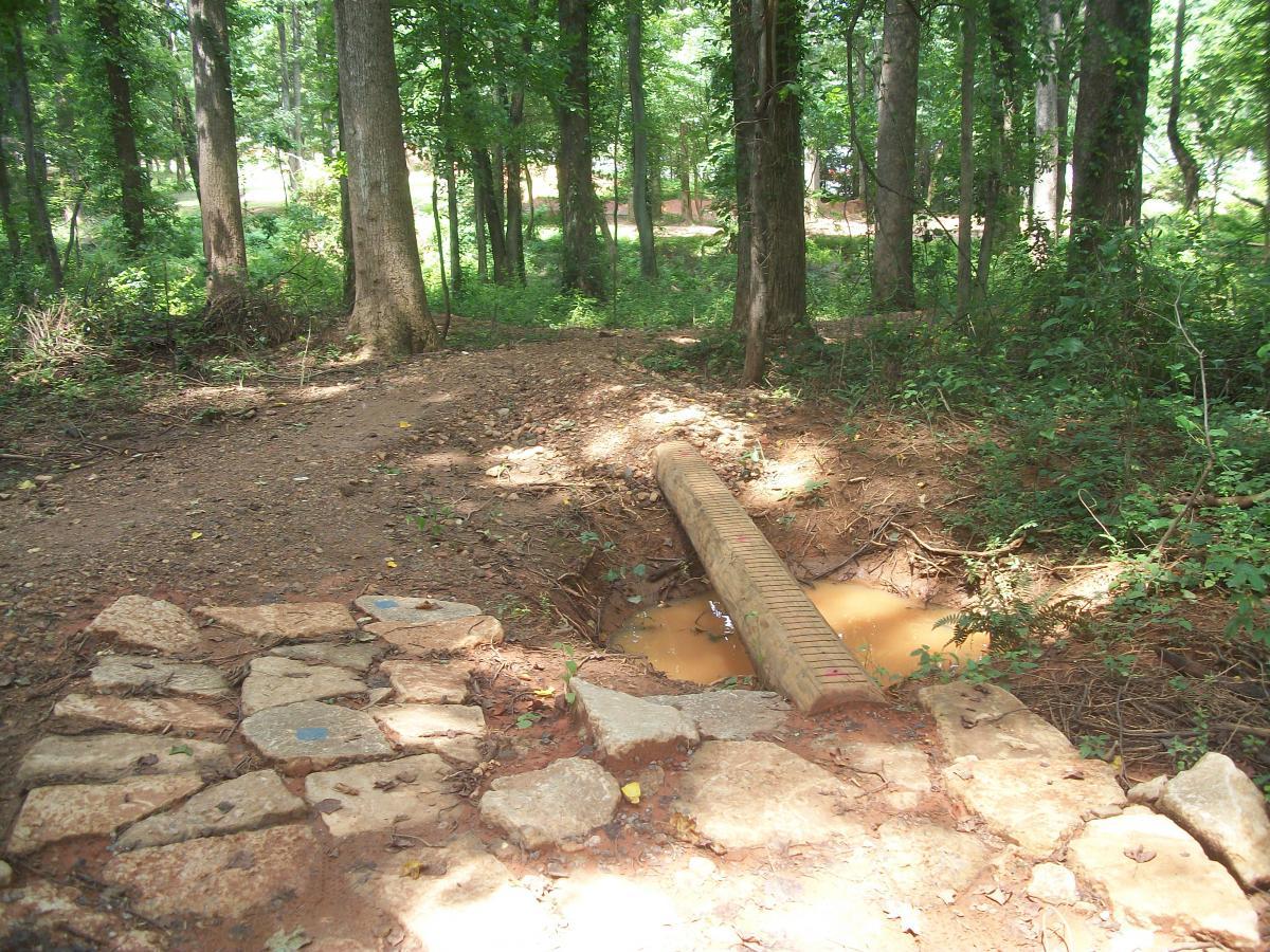 A dirt trail winding through a wooded area with tall trees and greenery. In the foreground, a stone path leads to a small drainage area with a rectangular concrete drain pipe crossing over muddy water. Sunlight filters through the leaves, casting dappled light on the ground. Gateway Mountain Bike Park mountain bike trail.