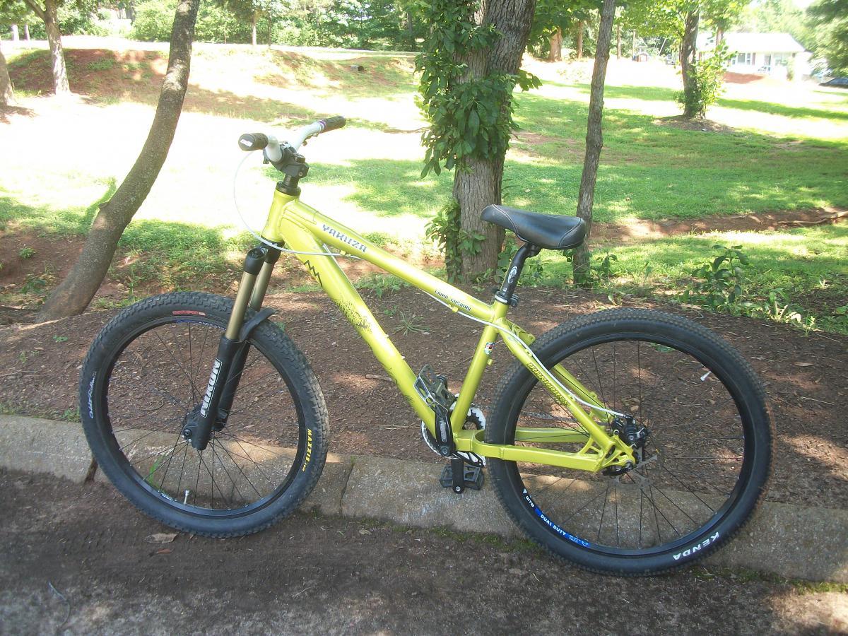 A bright yellow mountain bike parked on a gravel pathway near green trees and grass, featuring thick tires and a sturdy frame. Gateway Mountain Bike Park mountain bike trail.