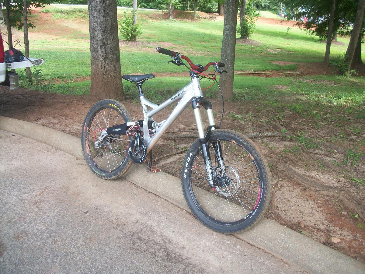 A silver mountain bike with black tires and red accents is leaning against a curb in a wooded area. The bike features a front suspension fork and has a dark black and red gear component visible. In the background, there are trees and a grassy area, suggesting a natural outdoor setting. Gateway Mountain Bike Park mountain bike trail.