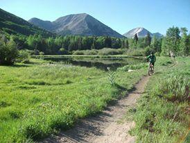 A scenic landscape featuring a dirt path alongside a serene lake, surrounded by lush greenery and mountains in the background under a clear blue sky. A person is biking on the path, enjoying the natural beauty of the outdoors. The Whole Enchilada mountain bike trail.