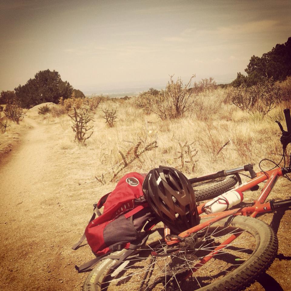 Kona Mahuna: A mountain bike resting on a dirt trail, with a red backpack and a black helmet placed on the ground. The scene features dry vegetation and shrubs surrounding the path, stretching into the distance under a clear sky.