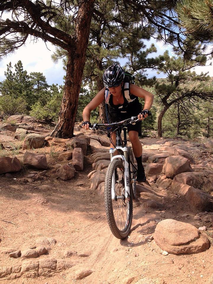 A cyclist wearing a helmet navigates a rocky trail surrounded by trees, showcasing a dynamic moment in mountain biking. The terrain consists of loose rocks and packed dirt, with a backdrop of green vegetation and a partly cloudy sky. Hall Ranch mountain bike trail.