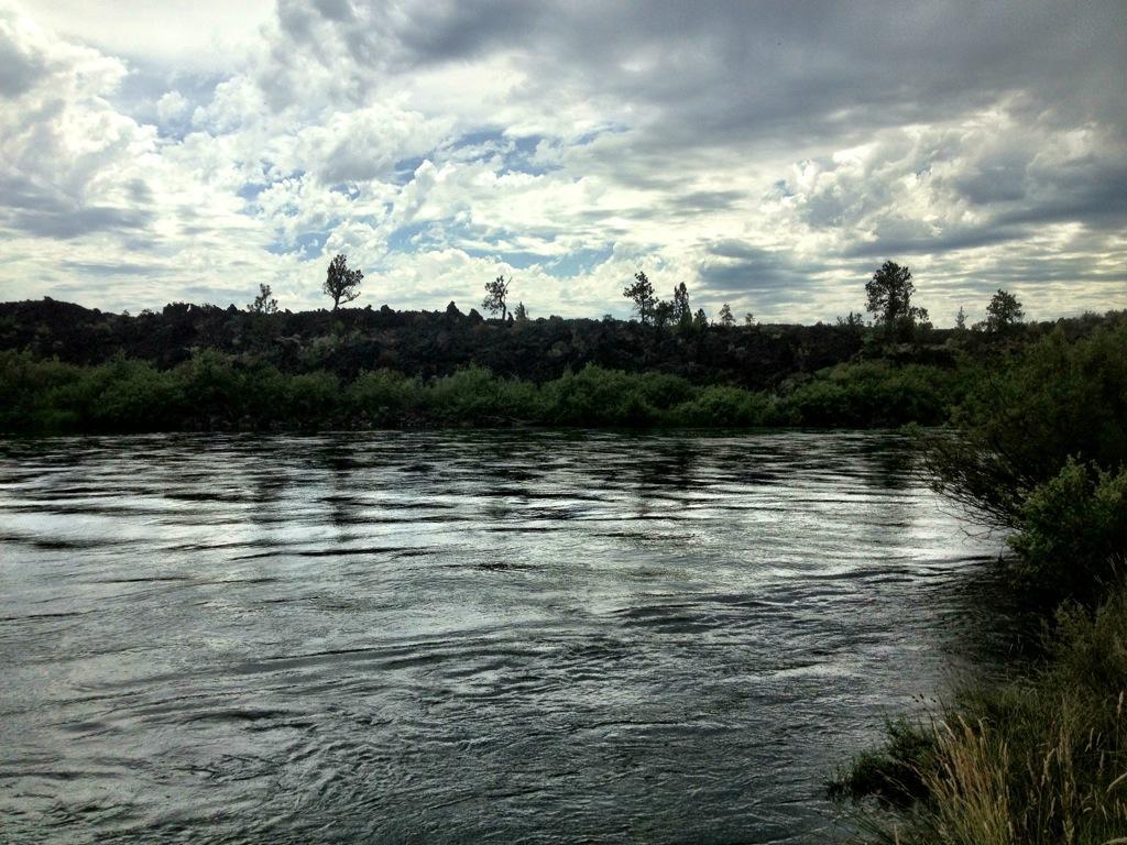 A serene river scene under a cloudy sky, featuring calm waters reflecting the sky and surrounded by lush greenery and rocky terrain in the background. Deschutes River mountain bike trail.