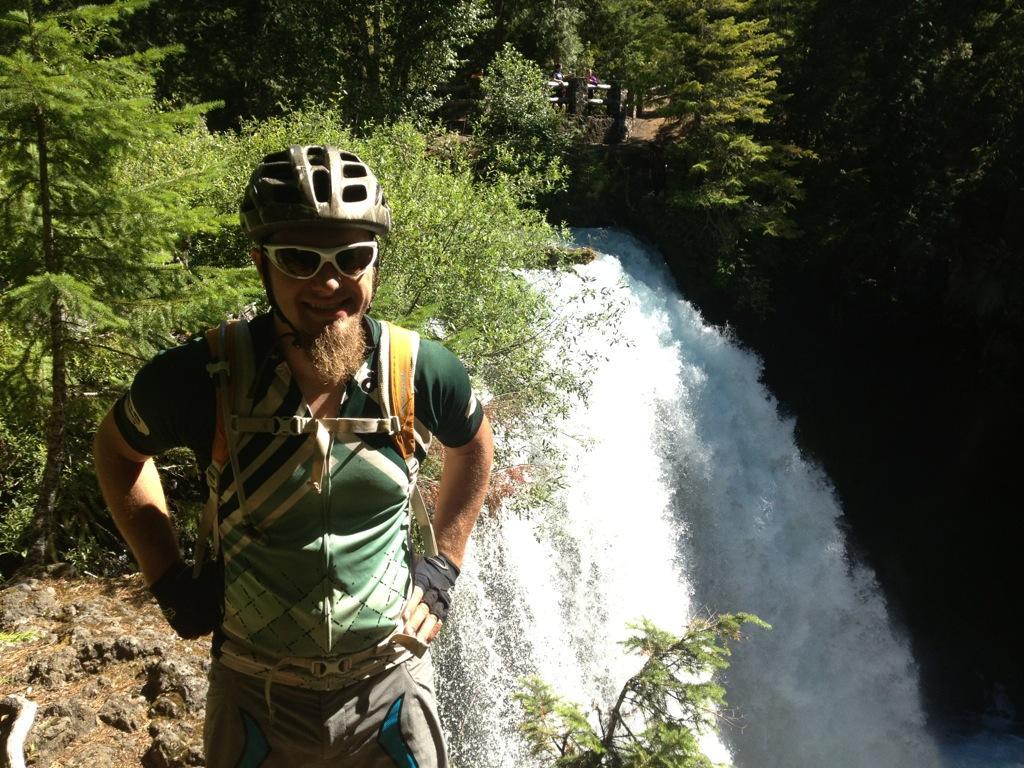 A person wearing a helmet and sunglasses stands in front of a waterfall, surrounded by green trees and vegetation. They are dressed in outdoor cycling attire, showcasing a playful pose with hands on hips, while water cascades down behind them. Mckenzie River Trail mountain bike trail.