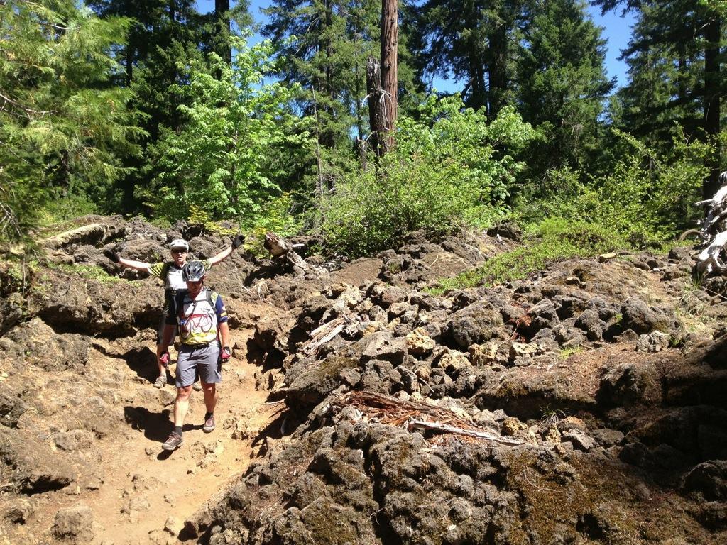 Two mountain bikers navigating a rocky trail surrounded by lush green trees and vegetation on a sunny day. One biker is in the foreground, focused on the path, while the other is behind, playfully posing with arms outstretched. Mckenzie River Trail mountain bike trail.