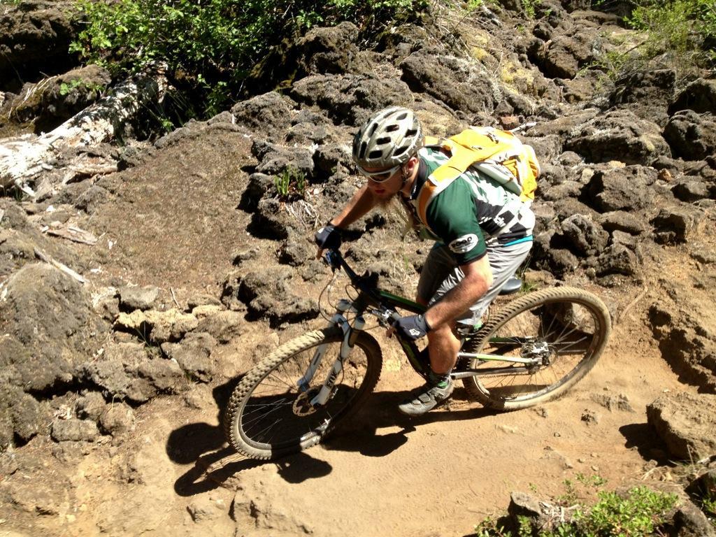 A person biking on a rocky trail surrounded by greenery, wearing a helmet and a backpack. The cyclist is navigating a challenging terrain with a focus on maintaining balance on the mountain bike. Mckenzie River Trail mountain bike trail.