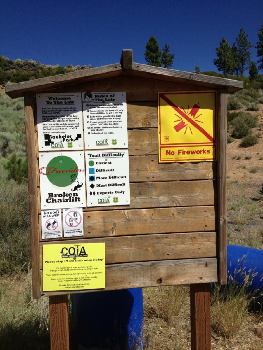 A wooden signpost displaying various informational notices, including trail rules and difficulty ratings for a recreational area. The sign features warnings against fireworks and dogs, along with a note about staying off muddy trails. In the background, there are trees and a clear blue sky, indicating an outdoor setting. The Lair mountain bike trail.