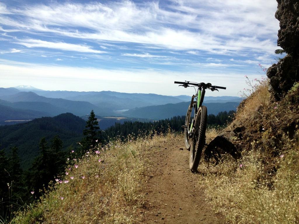 A mountain bike positioned on a dirt path at the edge of a cliff, overlooking a scenic view of rolling hills and mountains under a blue sky with wispy clouds. Wildflowers and grass line the trail, adding color to the landscape. Alpine Trail mountain bike trail.