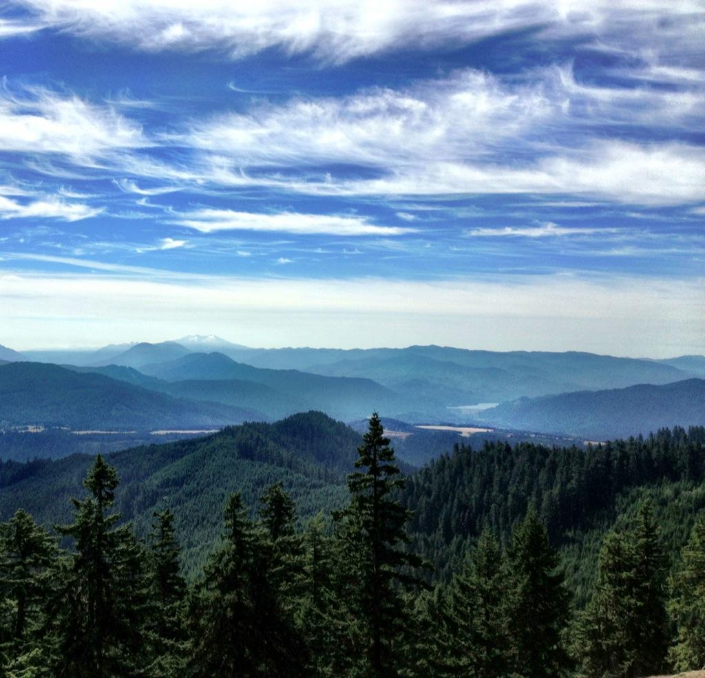 A panoramic view of a mountainous landscape featuring rolling green hills covered in evergreen trees, with a backdrop of distant mountains beneath a blue sky adorned with wispy clouds. The scene captures a serene and expansive natural environment. Alpine Trail mountain bike trail.