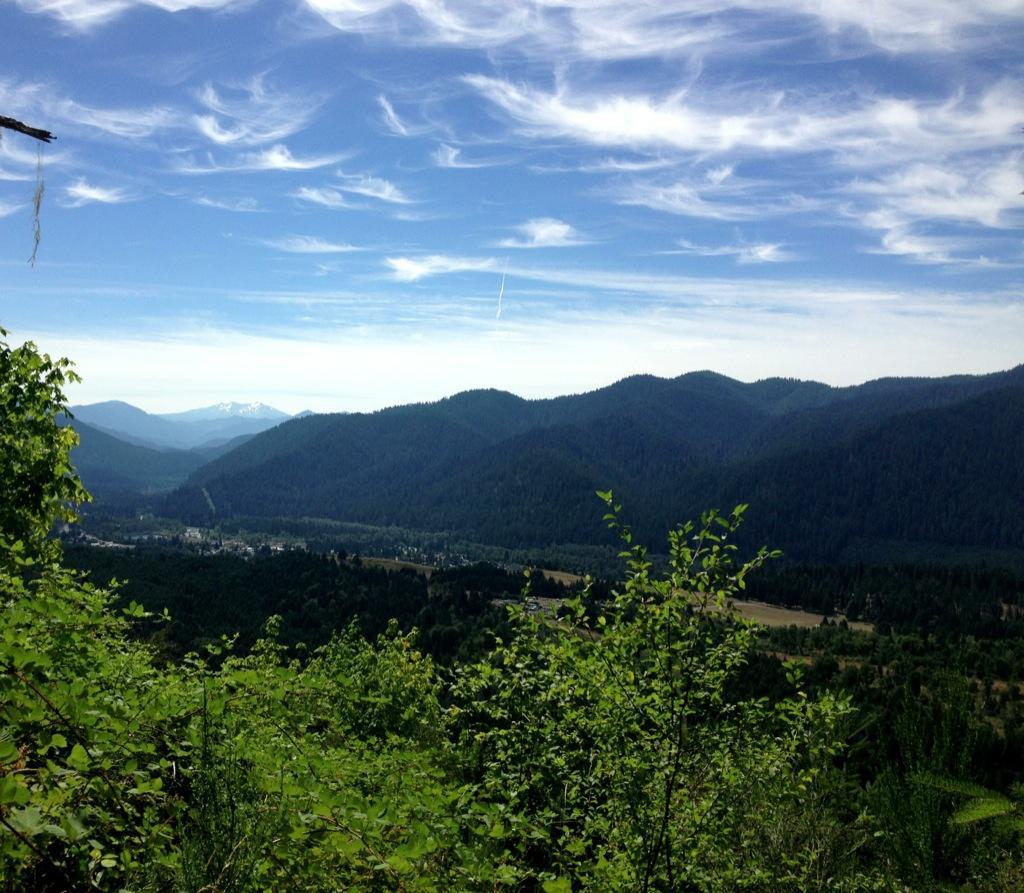 A panoramic view of rolling green mountains under a bright blue sky with wispy clouds. In the foreground, lush greenery frames the scene, while a small town is visible below, nestled in a valley. Snow-capped peaks can be seen in the distance, adding to the picturesque landscape. Alpine Trail mountain bike trail.