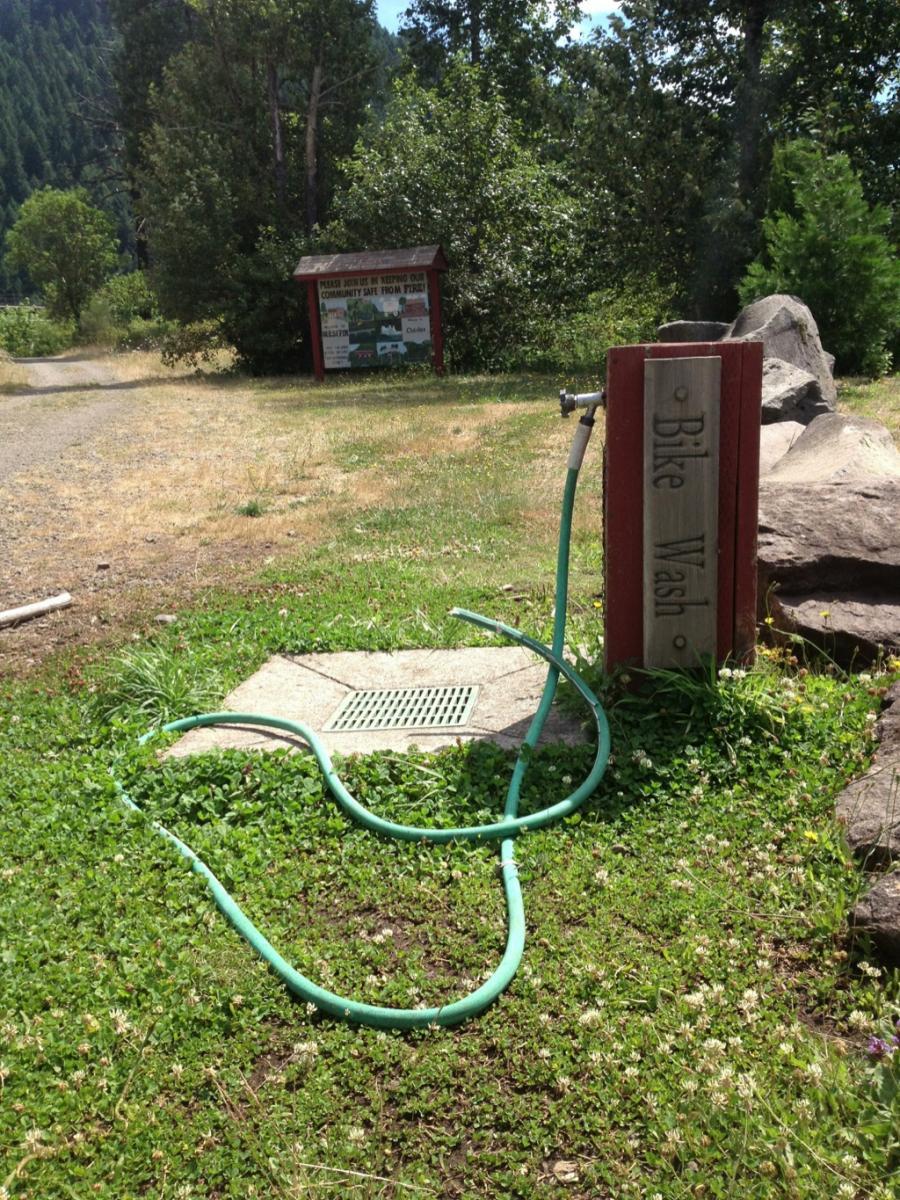A bike wash station featuring a green hose coiled on the ground next to a concrete pad with a drainage grate. In the background, there's a wooden signboard with informational posters, surrounded by trees and grassy areas under a clear sky. Alpine Trail mountain bike trail.