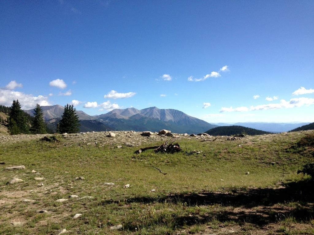 A scenic landscape featuring rolling hills and mountains under a blue sky with scattered clouds. The foreground shows a grassy area with rocks and fallen tree branches, while evergreen trees are visible on the left. In the background, the majestic mountain range stretches across the horizon. Monarch Crest Trail mountain bike trail.