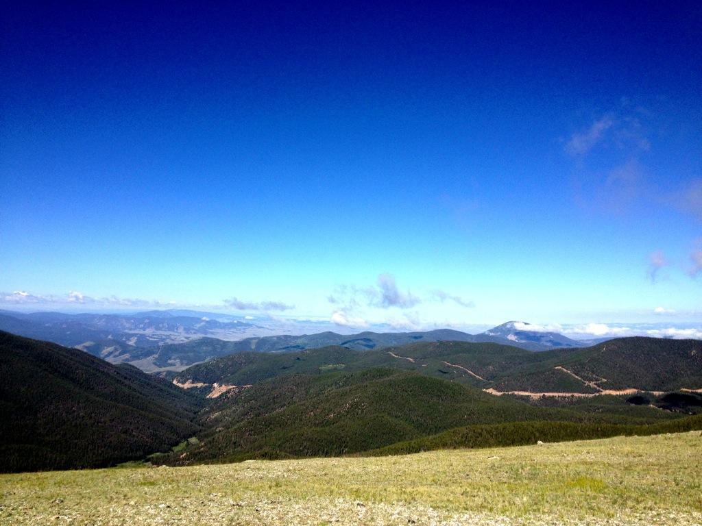 A panoramic view of rolling green mountains under a clear blue sky, with occasional clouds scattered across the horizon. The landscape features lush vegetation and distant mountain ranges fading into the background. Monarch Crest Trail mountain bike trail.