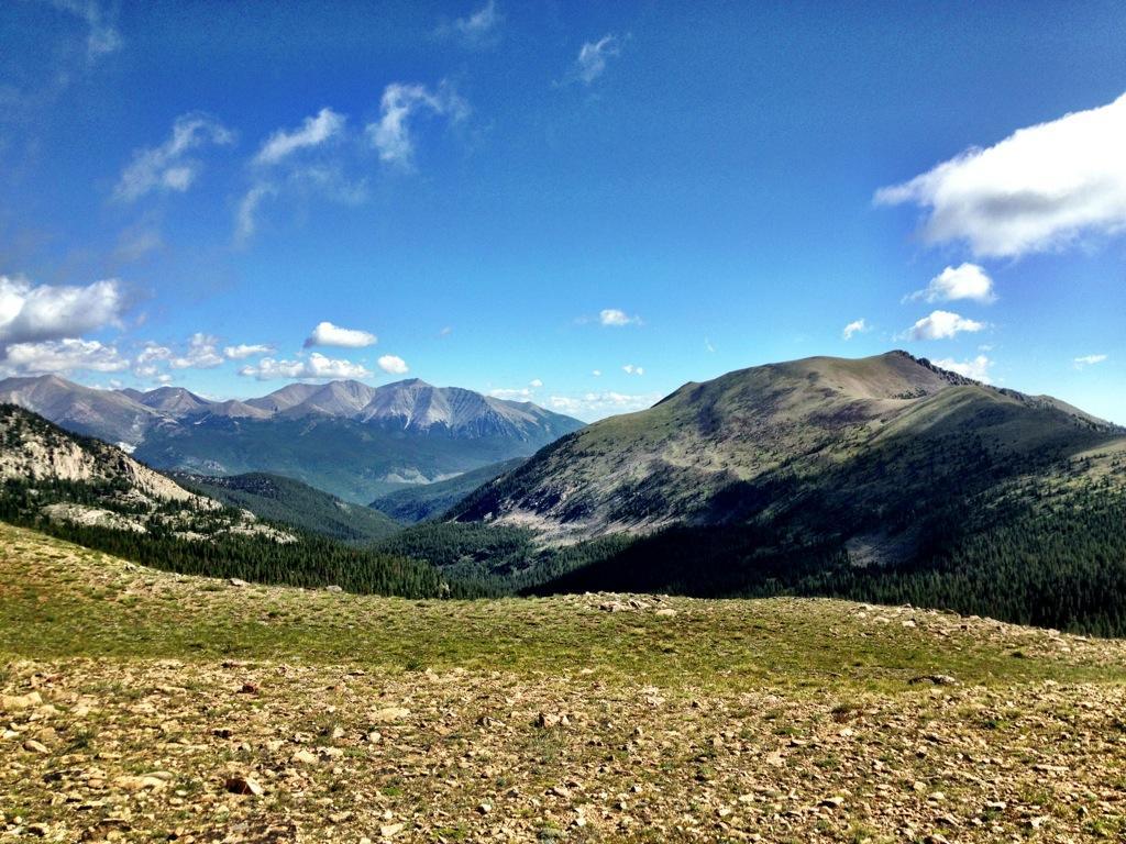 A panoramic view of rugged mountains under a clear blue sky, with rocky terrain in the foreground and lush greenery interspersed throughout the valleys. The distant peaks are partly covered in shadows, showcasing a variety of geological formations. Fluffy white clouds drift lazily across the sky, adding to the serene, natural beauty of the landscape. Monarch Crest Trail mountain bike trail.