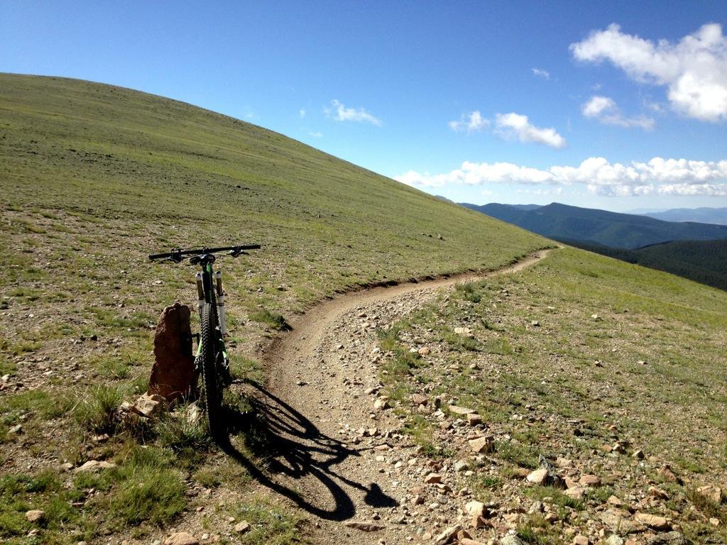 A mountain bike resting against a rocky outcrop on a winding dirt trail, surrounded by green hills under a clear blue sky with scattered clouds. Monarch Crest Trail mountain bike trail.