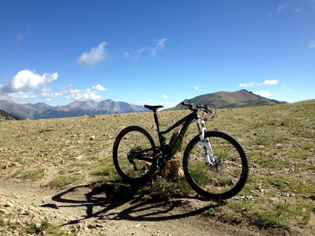 A mountain bike rests on a rocky trail with a scenic view of rolling hills and mountains under a clear blue sky, dotted with a few clouds. The ground is a mix of grass and gravel. Monarch Crest Trail mountain bike trail.
