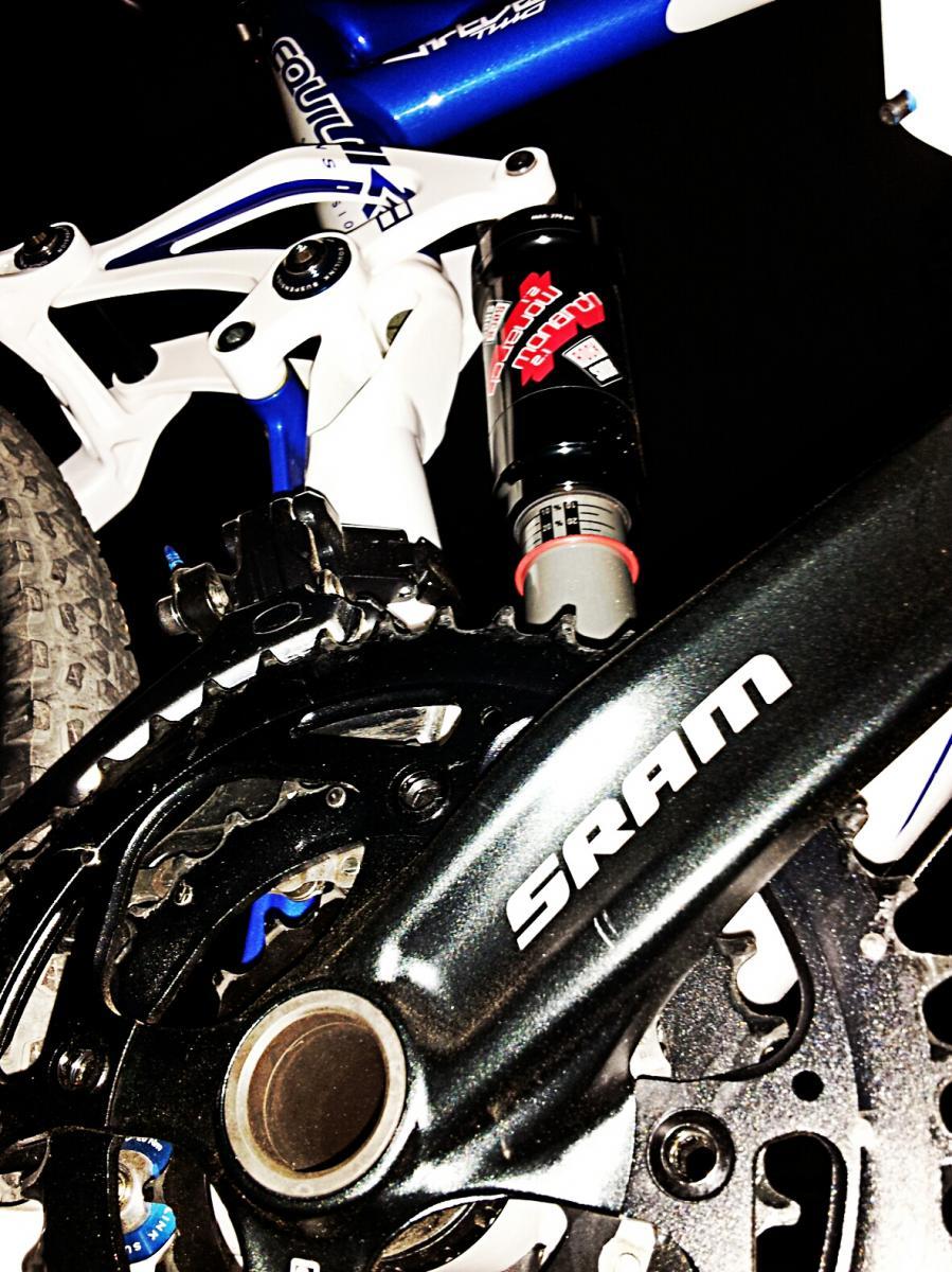 Felt Virtue Two: Close-up of a mountain bike's drivetrain, featuring a black SRAM crankset and chainring, along with part of the bike's suspension system in white and blue. The image highlights the bike's gear mechanism and the attached rear shock absorber.