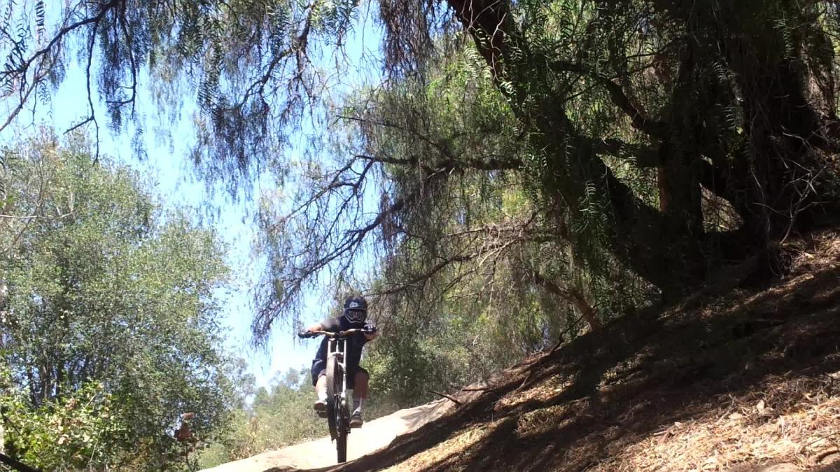 A person riding a mountain bike on a dirt trail in a wooded area, navigating a slope with trees and greenery surrounding them. The rider is slightly elevated, with the front wheel lifted off the ground, showcasing skill and adventure in an outdoor setting. Fullerton Loop mountain bike trail.