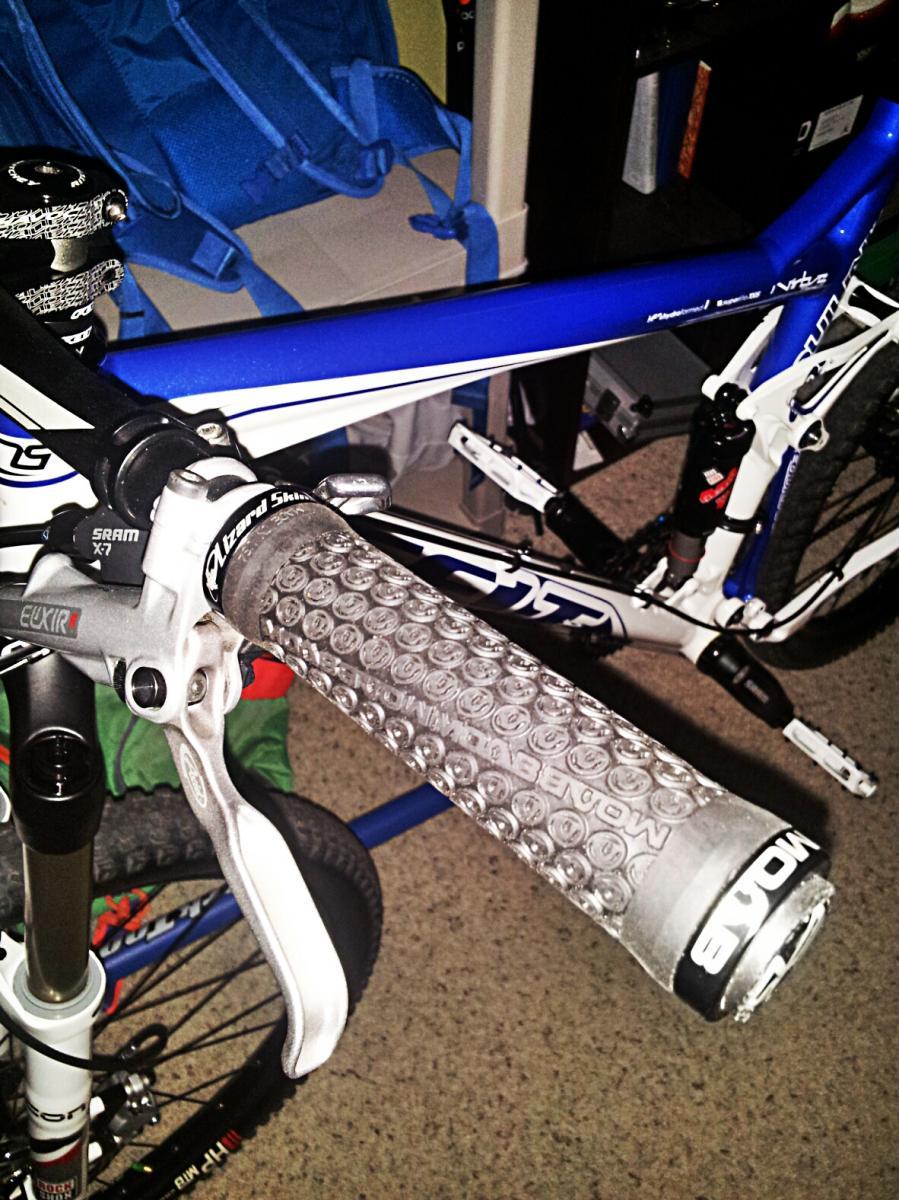Felt Virtue Two: Close-up image of a bicycle handlebar featuring a textured grip and a brake lever, showcasing part of a blue and white mountain bike in a room. The background includes a backpack and various items, with carpeted flooring visible.