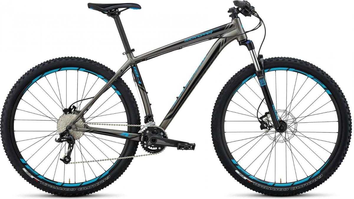 Specialized Rockhopper Comp 29: A modern mountain bike featuring a sleek gray frame with blue accents, wide tires designed for off-road terrain, and disc brakes. The bike is shown from a side angle, highlighting its sturdy construction and gear setup.