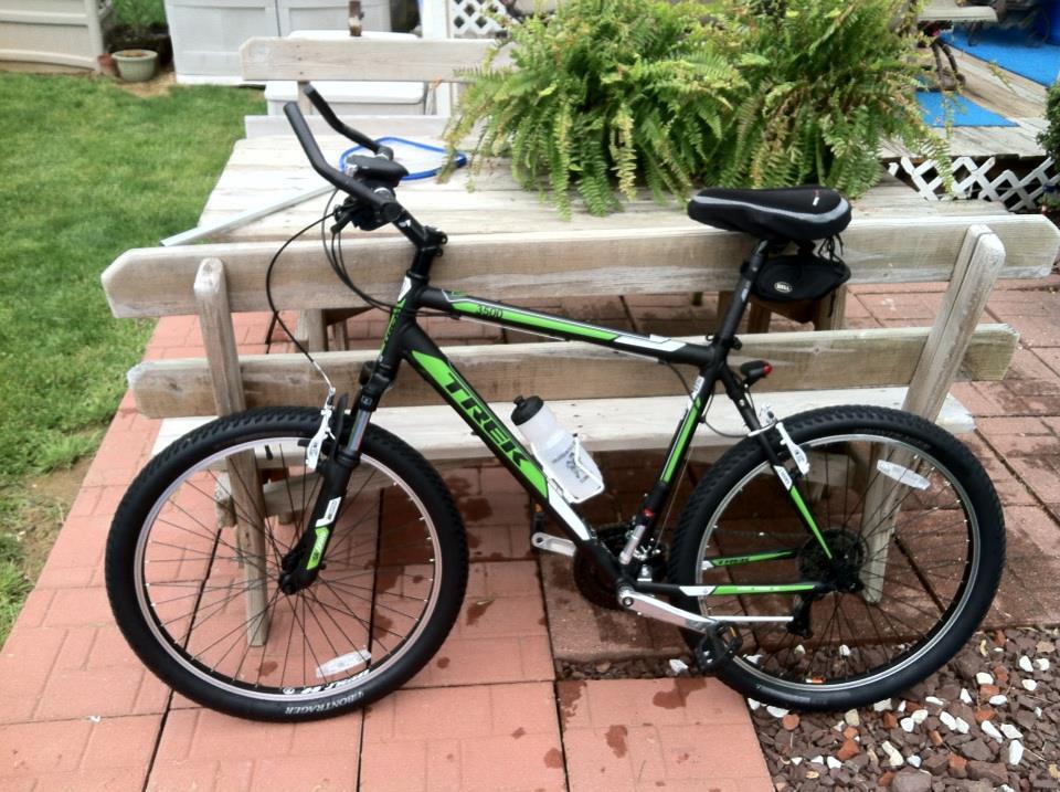 Trek 3500: A black and green Trek mountain bike is parked next to a wooden bench in a grassy outdoor area. The bike features knobby tires, a water bottle holder, and is positioned on a patio made of red bricks with some small pebbles scattered nearby. Lush green foliage is visible in the background.