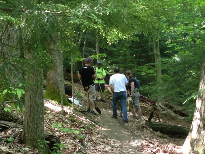A group of individuals gathered on a dirt trail in a wooded area, discussing plans or working on a project. Sunlight filters through the leaves, illuminating the scene, with trees and foliage surrounding them. Some participants are holding tools, and the atmosphere appears collaborative and outdoorsy.