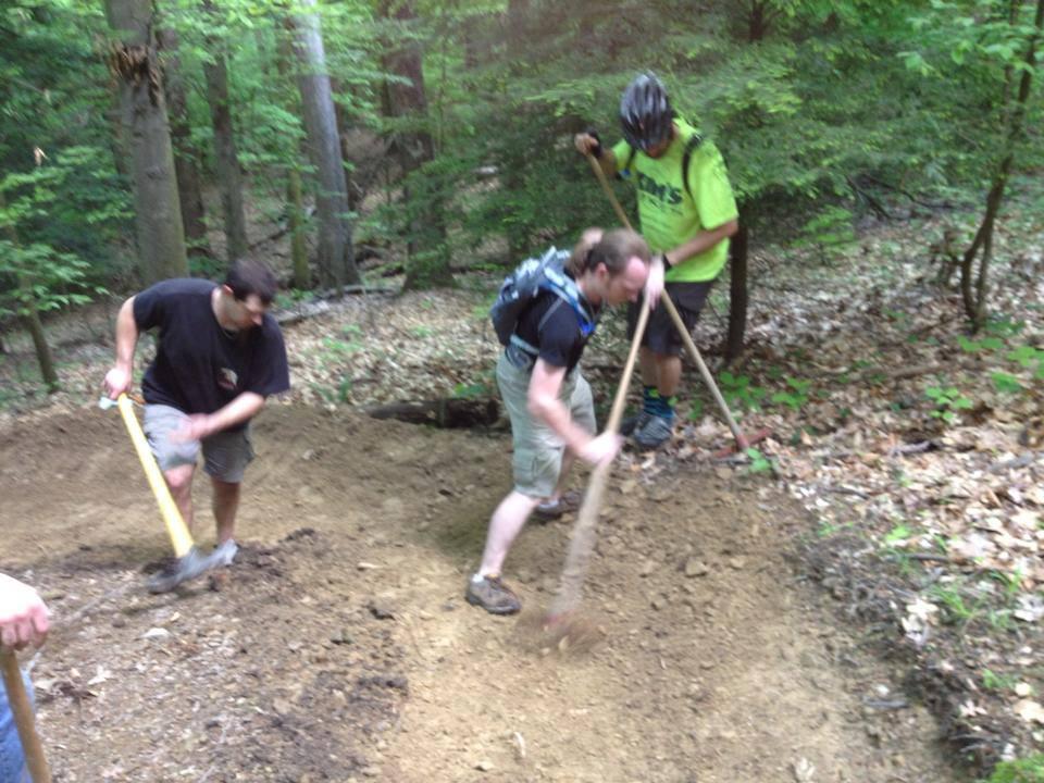 Three individuals working together to build or maintain a trail in a wooded area. They are using tools such as shovels and rakes, surrounded by trees and natural foliage. The scene conveys teamwork and outdoor activity.