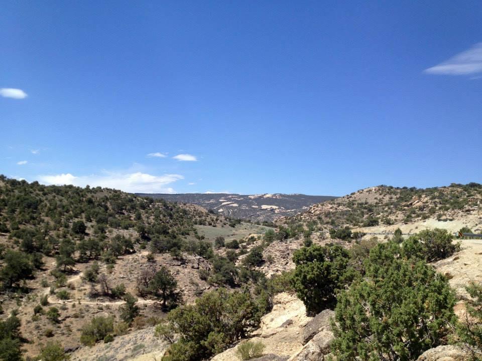 A panoramic view of rolling hills and rocky terrain under a clear blue sky, with scattered shrubs and trees dotting the landscape. The scene captures the natural beauty of a dry, mountainous region, showcasing the diverse textures of the earth. The Ribbon mountain bike trail.