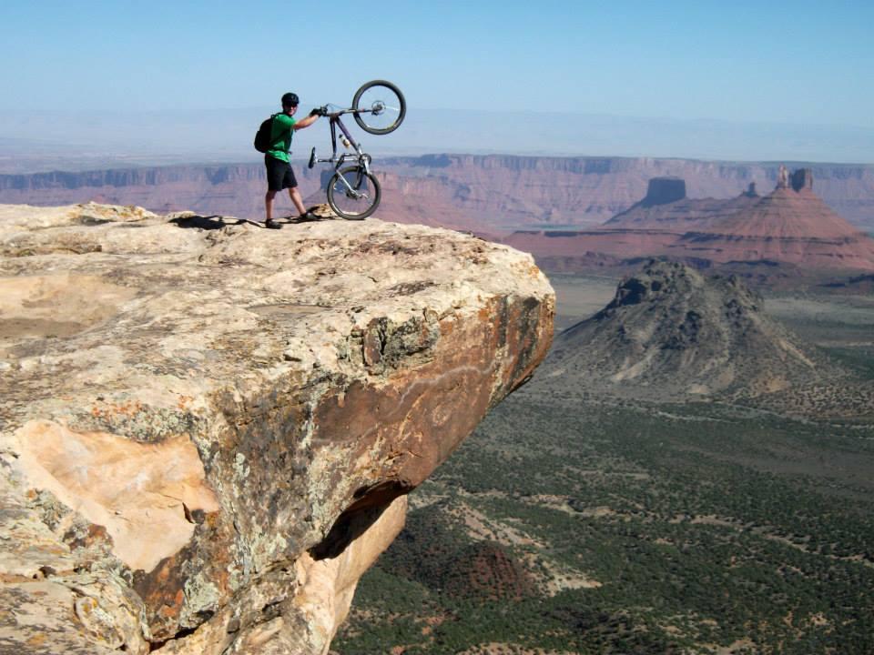 A person standing on the edge of a rocky cliff, holding a bicycle above their head. The scene features a wide landscape with distant mesas and mountains under a clear blue sky, illustrating a sense of adventure and breathtaking nature. The Whole Enchilada mountain bike trail.