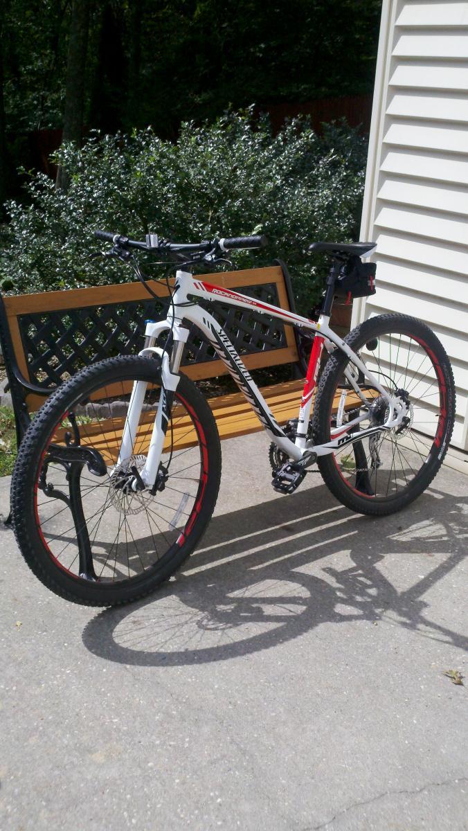 Specialized Hardrock Disc 29: A white mountain bike with red accents is parked on a paved surface next to a brown wooden bench. Green foliage is visible in the background, suggesting an outdoor setting. The bike features thick tires, a front suspension, and visible gear components. Shadowing from the bike creates an interesting pattern on the ground.
