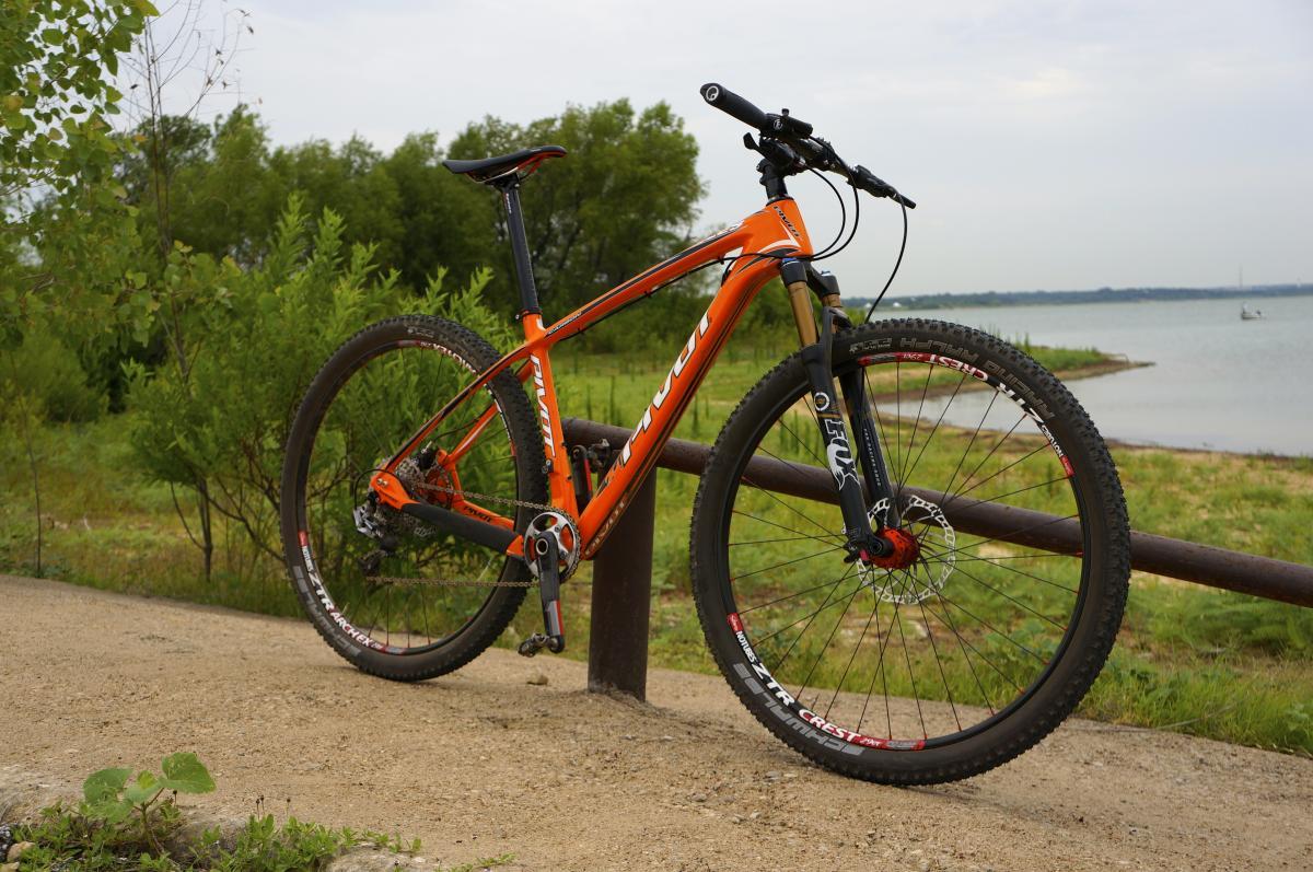 Pivot Les: A bright orange mountain bike is leaning against a wooden railing along a path near a body of water. Lush green trees and grass surround the area, providing a natural backdrop. The sky is overcast, suggesting a calm day.