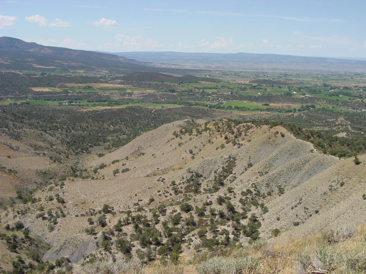 A panoramic view of rolling hills and valleys, showcasing a landscape with patches of green fields and sparse vegetation. The scene is under a clear blue sky with a few scattered clouds, with distant mountains visible on the horizon. Jumbo Mountain mountain bike trail.