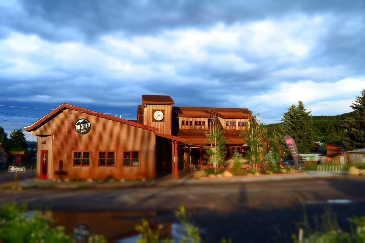 A rustic building with a corrugated metal exterior featuring large windows and a clock on the front. The structure has a sloped roof and is surrounded by green trees and landscaped areas. A sign indicates the name "Tin Shed." The sky is overcast, adding a dramatic atmosphere to the scene.