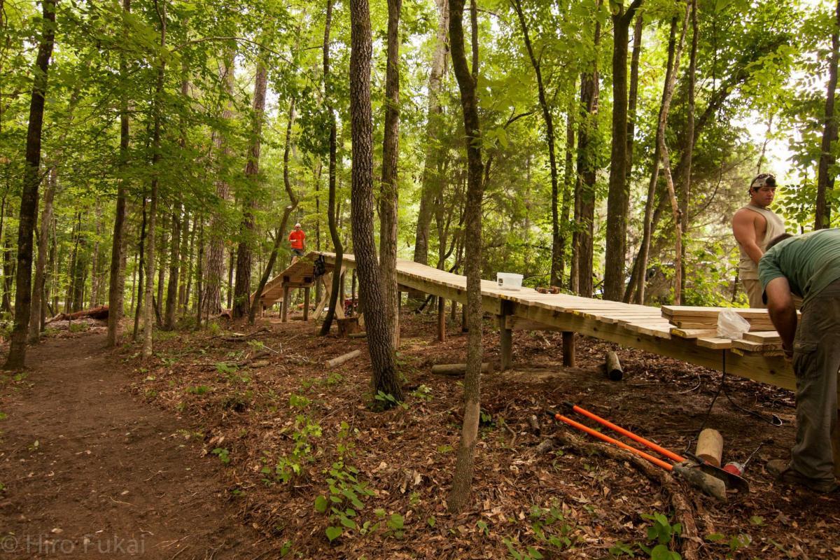 Construction of a wooden bridge is taking place in a dense forest. Two individuals are working on the bridge, with one person wearing an orange shirt standing on the structure, while another is kneeling beside wooden planks and tools on the ground. The scene is surrounded by tall trees and green foliage, showcasing the natural environment. The path in the foreground is clear, leading into the woods. Chewacla State Park mountain bike trail.