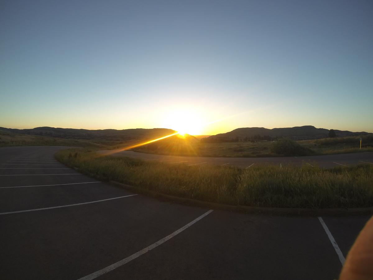 Fuji Nevada 1.0: Sunrise over rolling hills with a clear sky, viewed from a parking area with grass and empty parking spaces in the foreground. The sun casts a warm glow and creates a lens flare effect as it rises on the horizon.