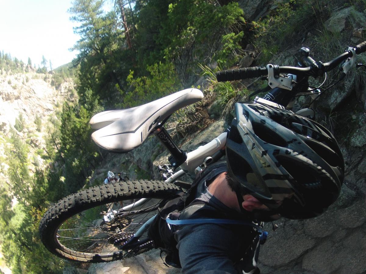 A mountain biker navigating a rocky trail, holding up their bike at a steep angle. The background features lush green trees and rugged terrain, suggesting an outdoor adventure in a natural setting. The cyclist is wearing a helmet, emphasizing safety while biking. Walker Ranch mountain bike trail.