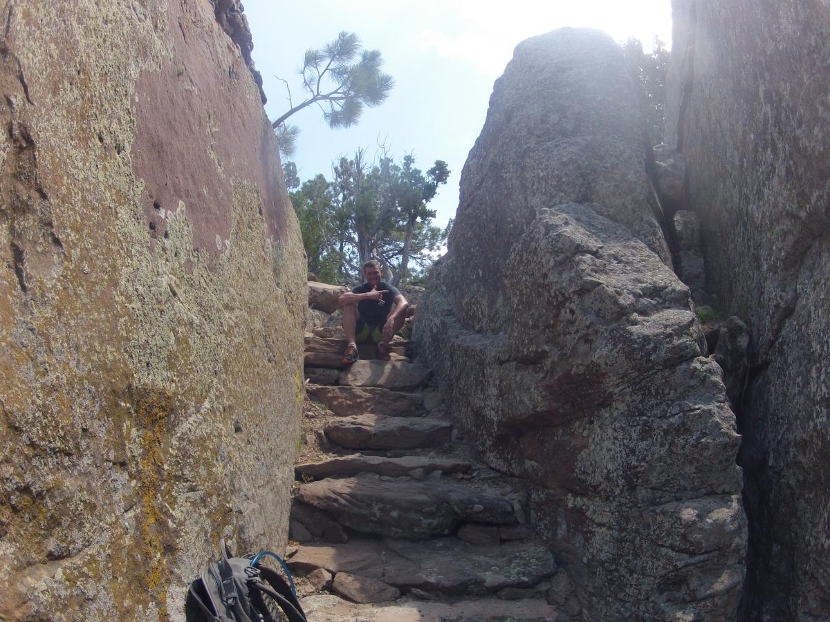 A person sitting on a rocky staircase between two large stone formations, surrounded by a natural landscape with trees in the background. The stones are textured with moss and lichen, and the scene is illuminated by bright sunlight. A backpack is positioned nearby on the ground. Glendo State Park mountain bike trail.
