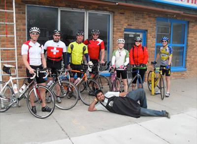A group of seven cyclists in colorful biking gear, standing with their bicycles outside a brick building. One person in an apron is lying on the ground in front of them, smiling. A ladder is positioned nearby.