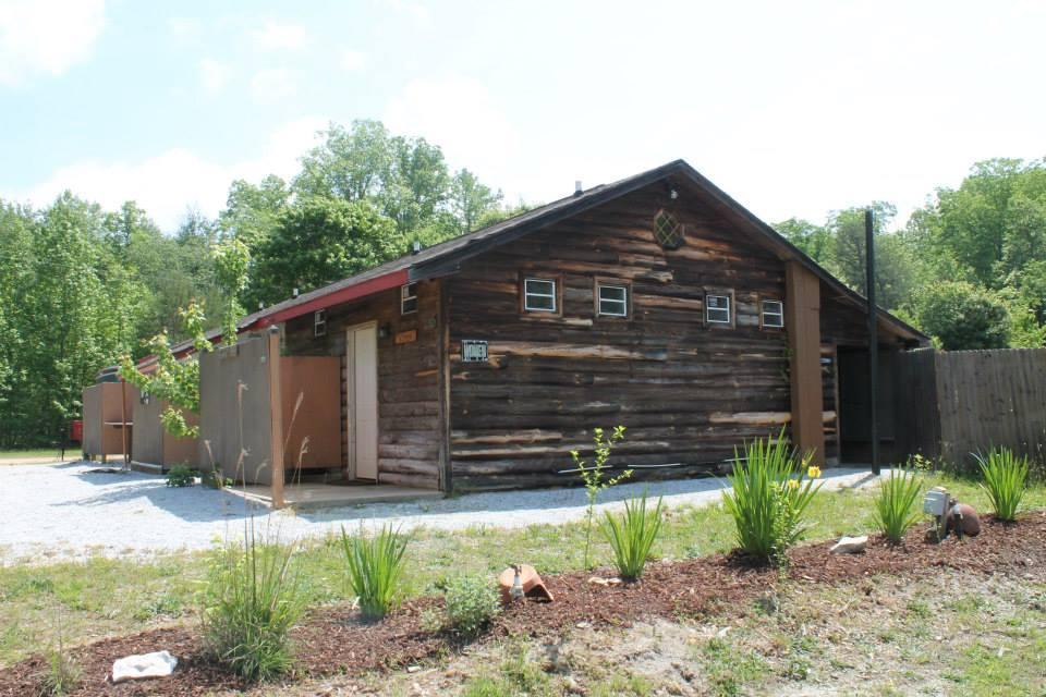 A rustic wooden cabin with a sloped roof, set against a backdrop of green trees. The cabin features multiple small windows and a door, with a gravel driveway in front and landscaped greenery, including small plants and decorative stones, surrounding the structure. Hilltop Bike Park mountain bike trail.